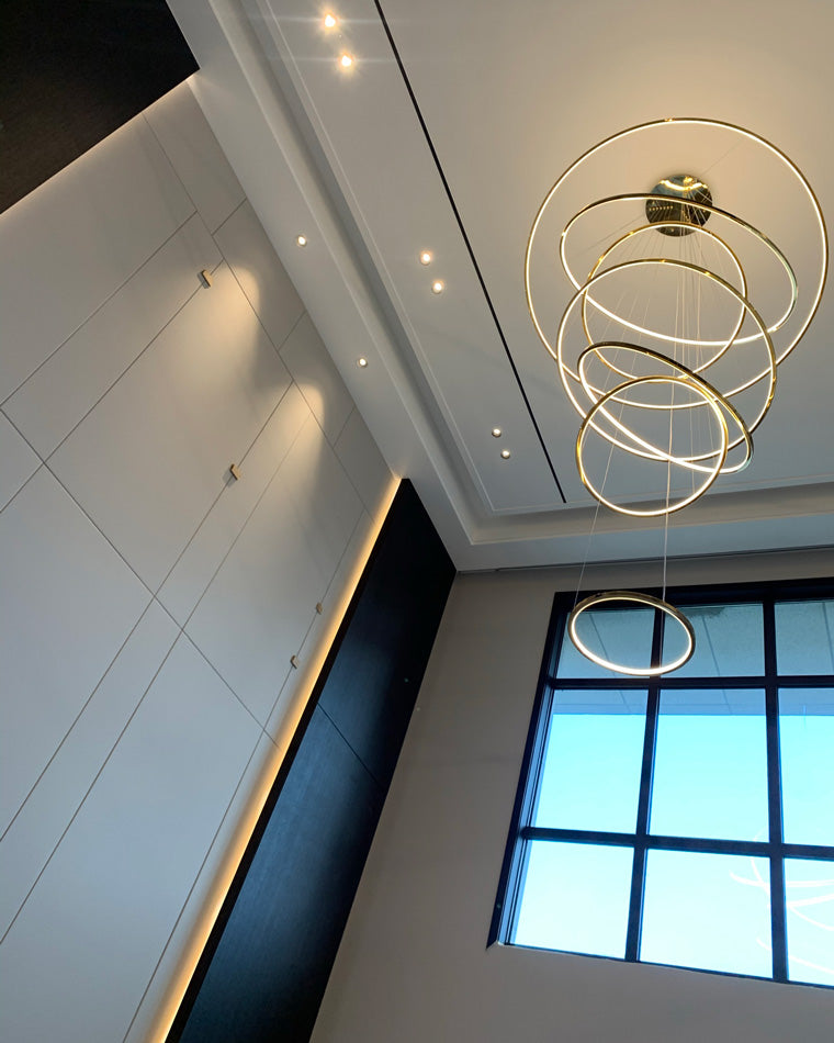 Golden LED rings glowing beneath a tall ceiling, emphasizing the chandelier’s airy cables and dramatic vertical reach.