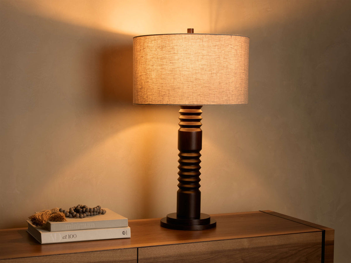 Front view of a dark brown carved base table lamp with a cylindrical light beige fabric shade, illuminated.