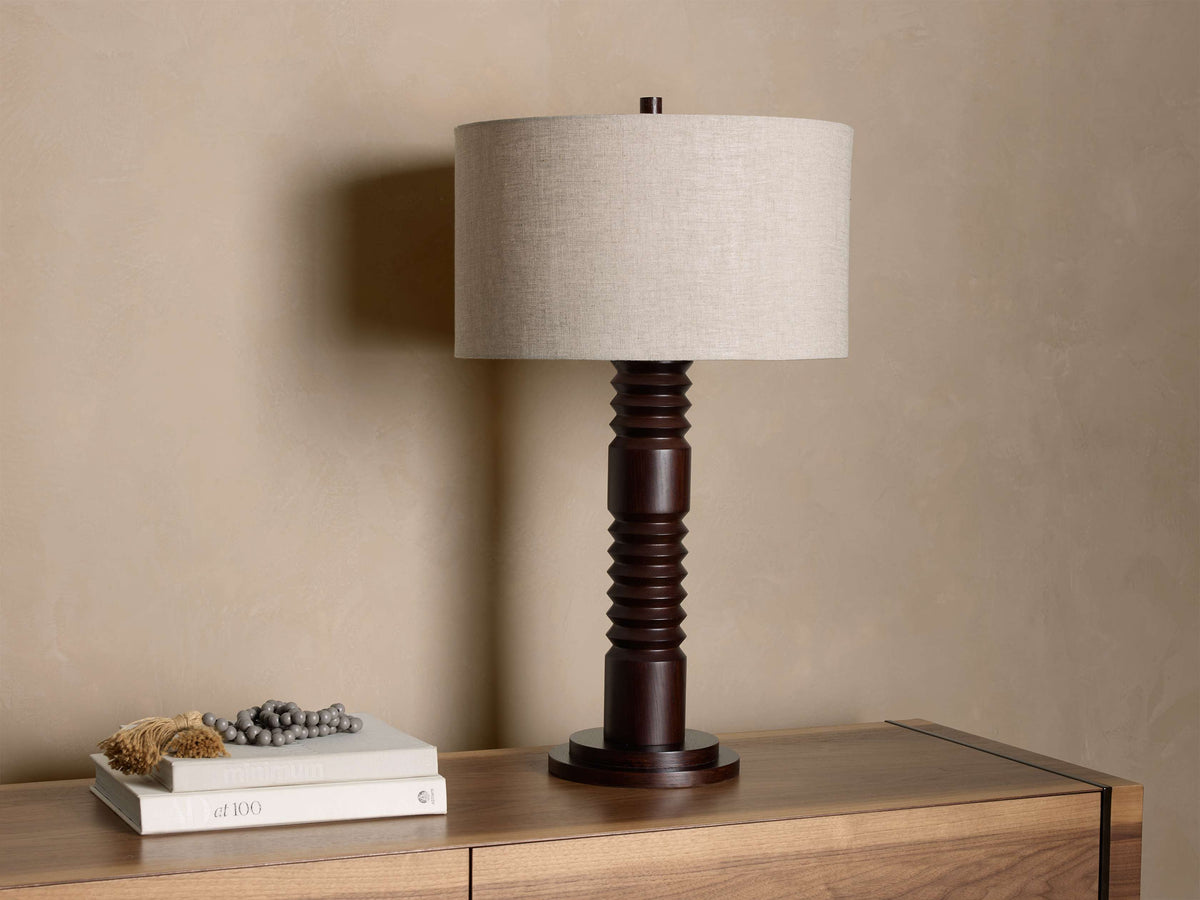 Front view of a dark brown carved wood table lamp with a light beige linen cylindrical shade.