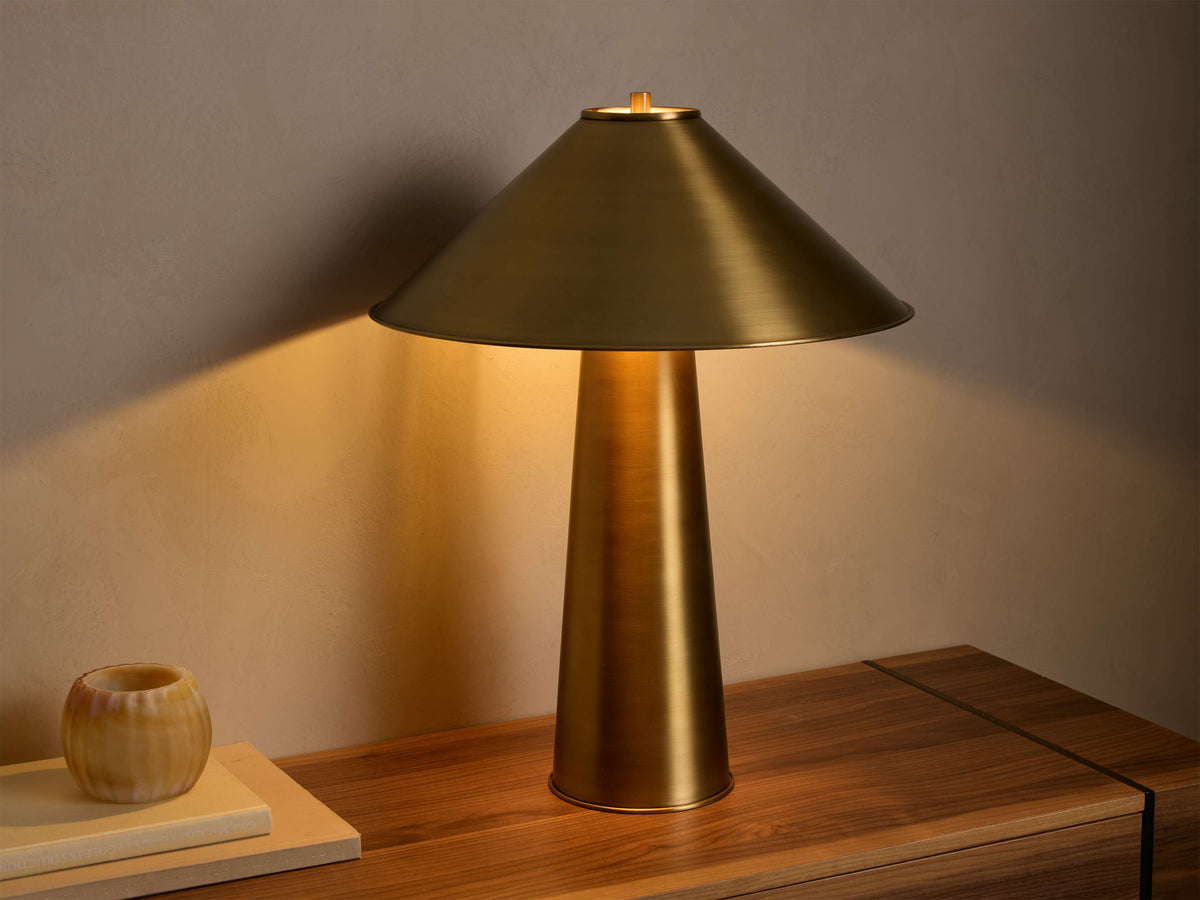 Lit antiqued brass Ombra Metal Table Lamp, featuring a conical shade and cylindrical base, casting warm light on a wooden surface.