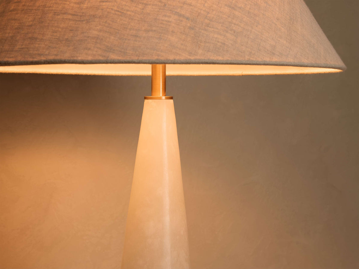 Close-up of a table lamp with a gray fabric lampshade and a gold-toned stem over a light-colored conical base, emitting warm light.