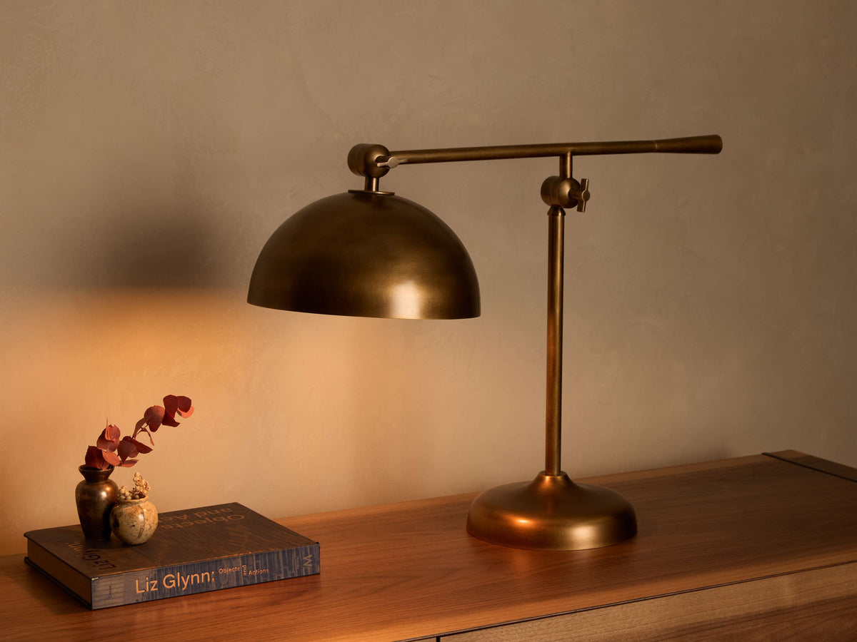 Front view of a brass table lamp with a dome shade, adjustable arm, and round base, casting a warm light on a wooden desk.