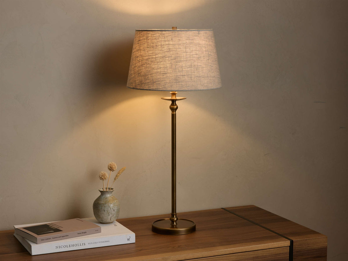 Front view of the Winslow Table Lamp with an antique brass base and light grey fabric shade, illuminated, on a wooden table.