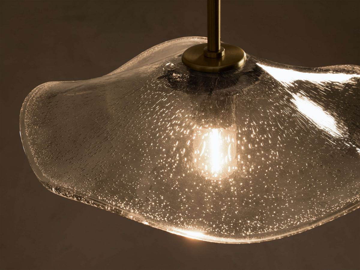 Close-up of a clear seeded glass wavy pendant light with a warm glowing bulb, suspended by a brass rod.