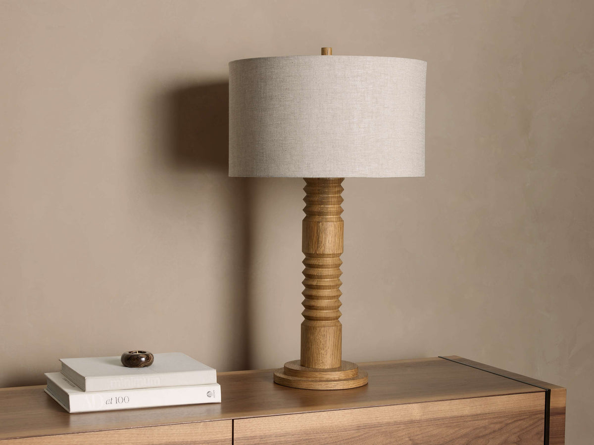 Front view of a Carved Wood Table Lamp with a natural wood spindle base and a light gray fabric drum shade, sitting on a wooden console.