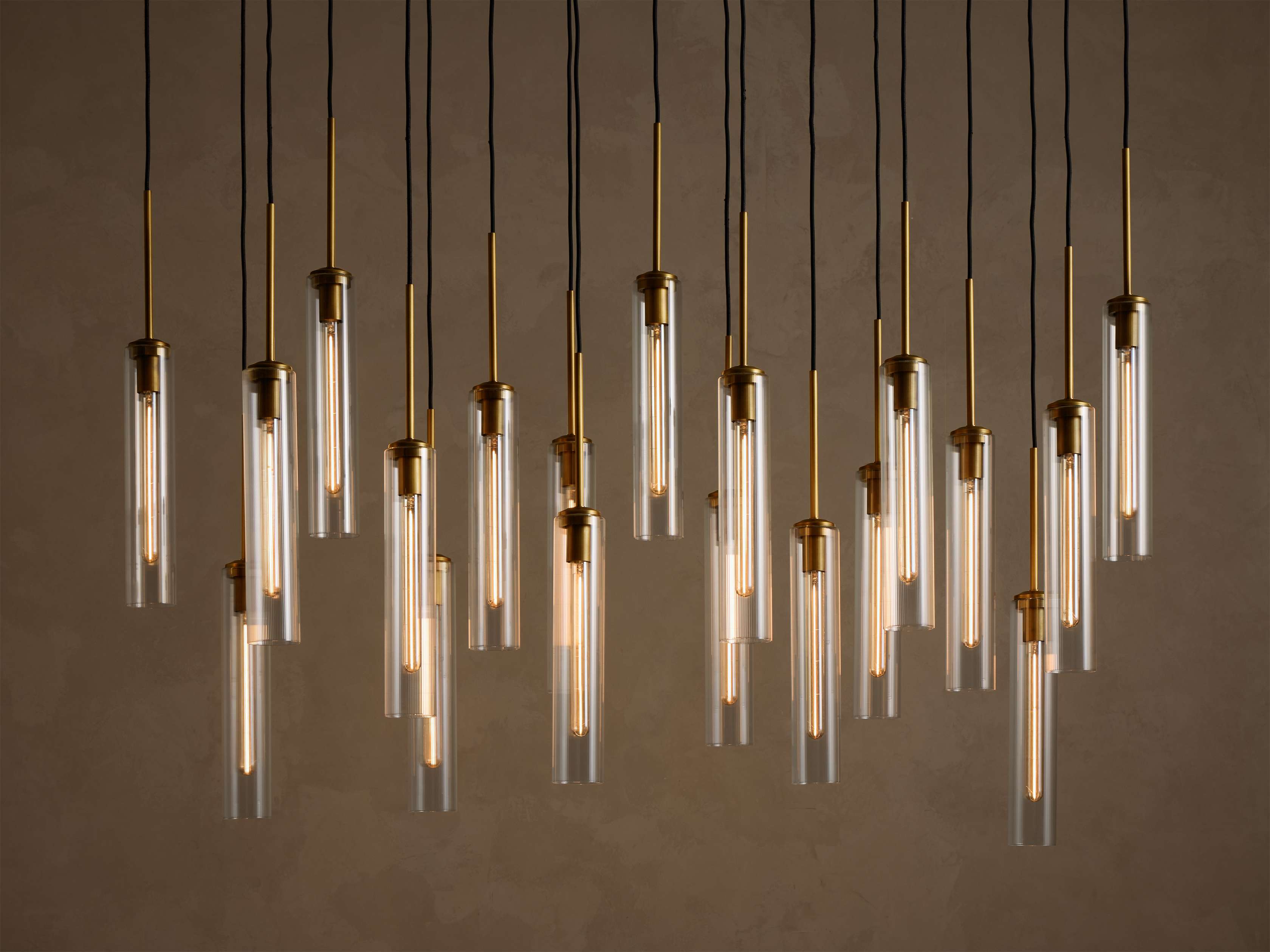 Multiple illuminated pendant lights with clear glass tubes and brass fixtures, hanging at varying heights from black cords.
