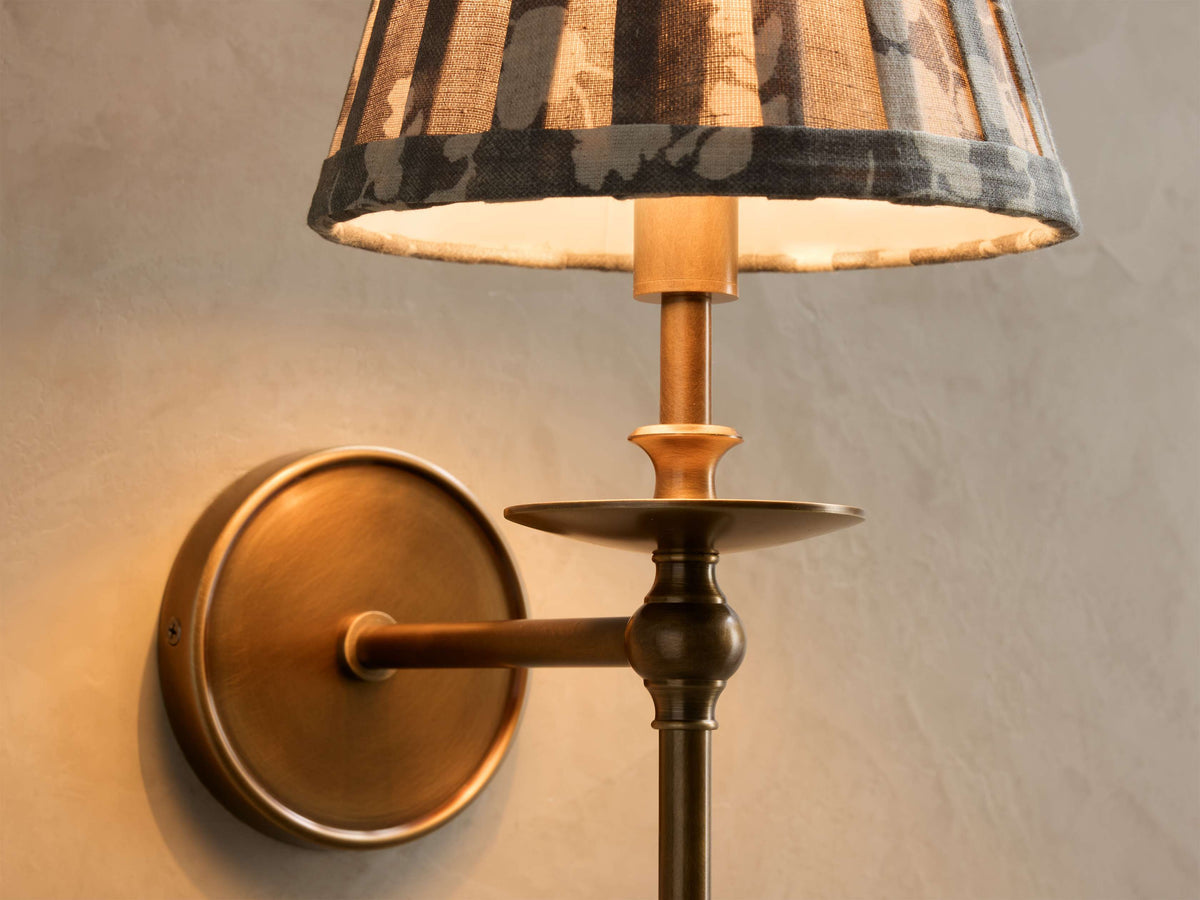 Close-up of the antique brass Winslow Single Sconce with a pleated grey and cream floral fabric shade, lit.