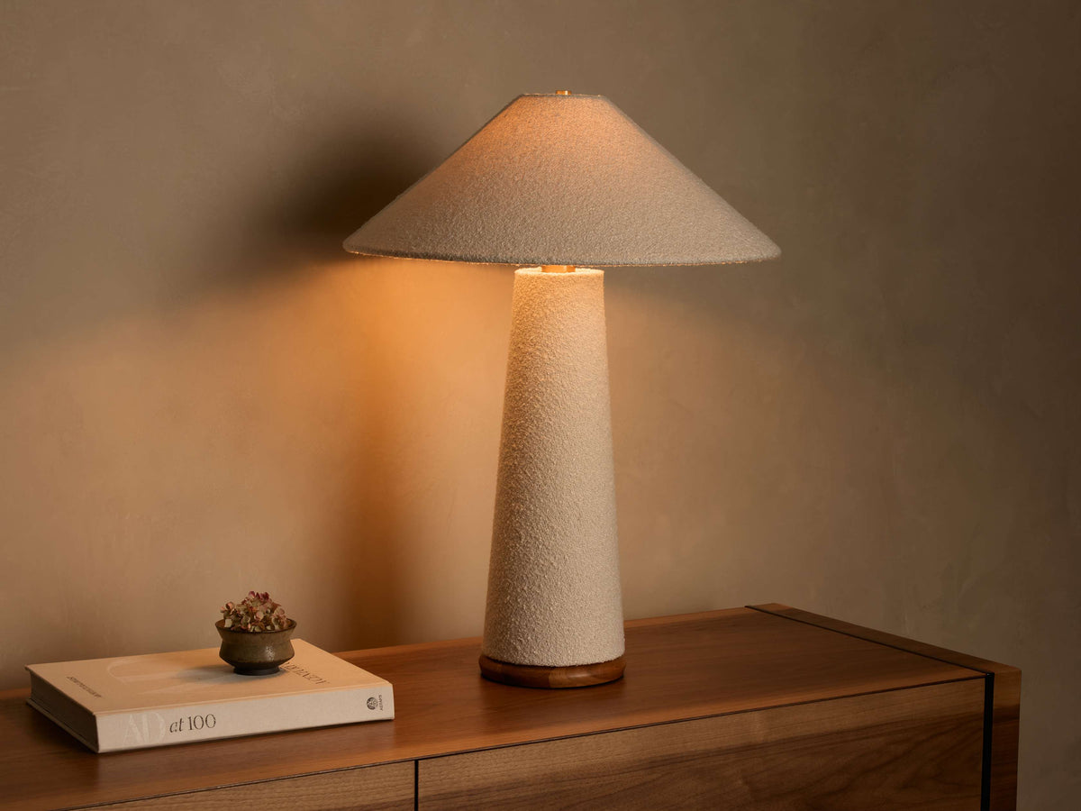 Front view of a lit table lamp featuring a light beige textured cone shade and matching tapered cylindrical base, on a wooden surface.