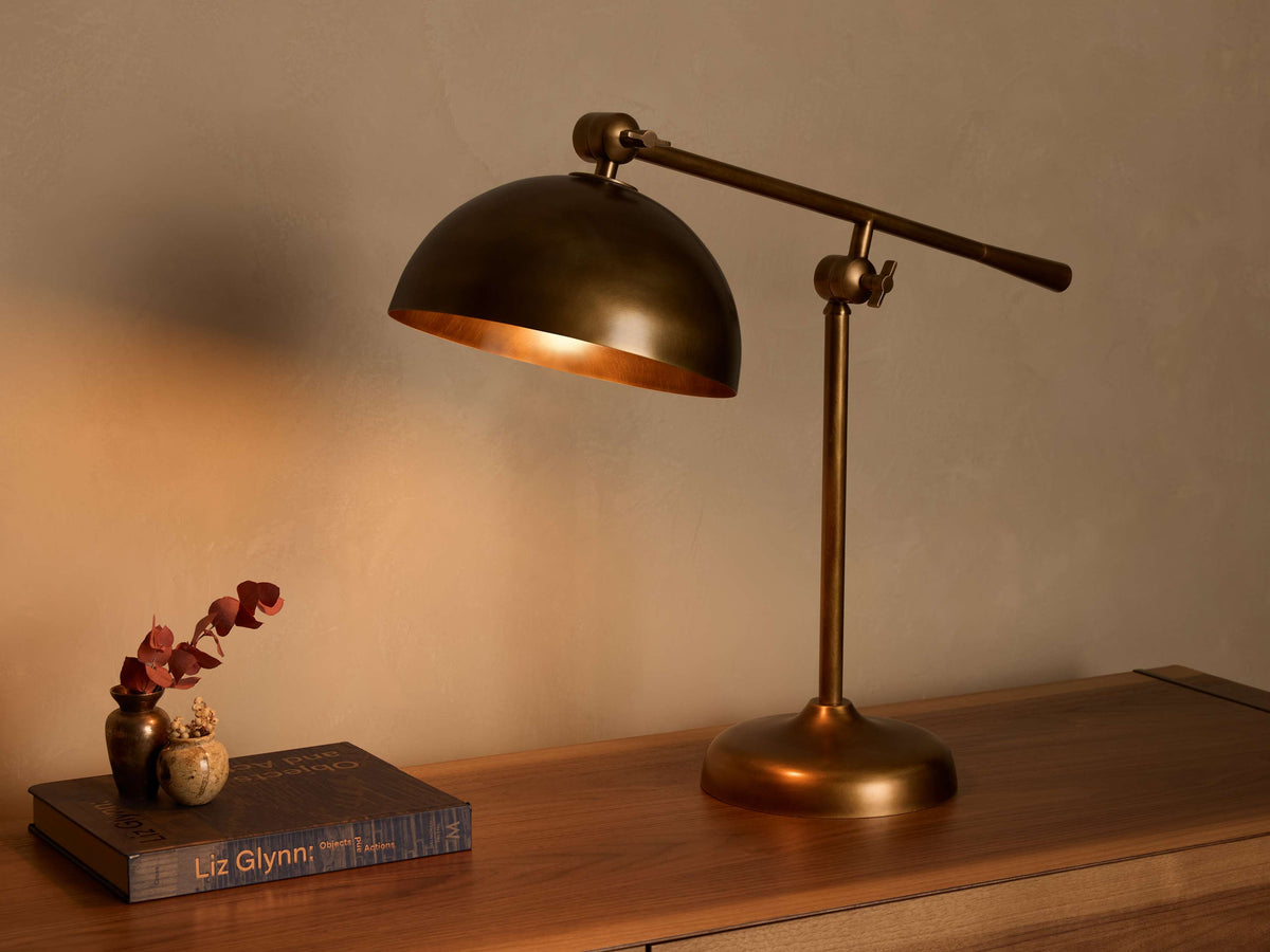 Front view of an illuminated brass industrial-style adjustable table lamp with a domed shade on a wooden surface.