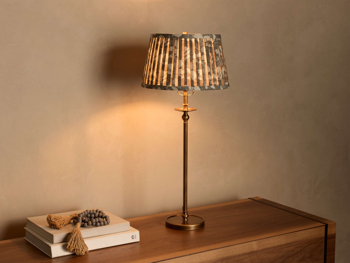 Front view of an illuminated Winslow Table Lamp with a dark pleated fabric shade and an antique brass base, on a wooden dresser.