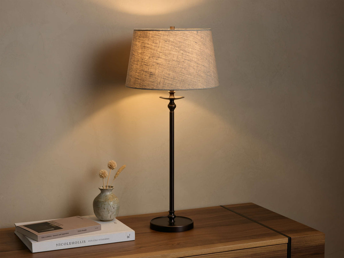 Front view of the Winslow Table Lamp, with a light natural fabric shade and dark bronze iron base, illuminated on a wooden table.