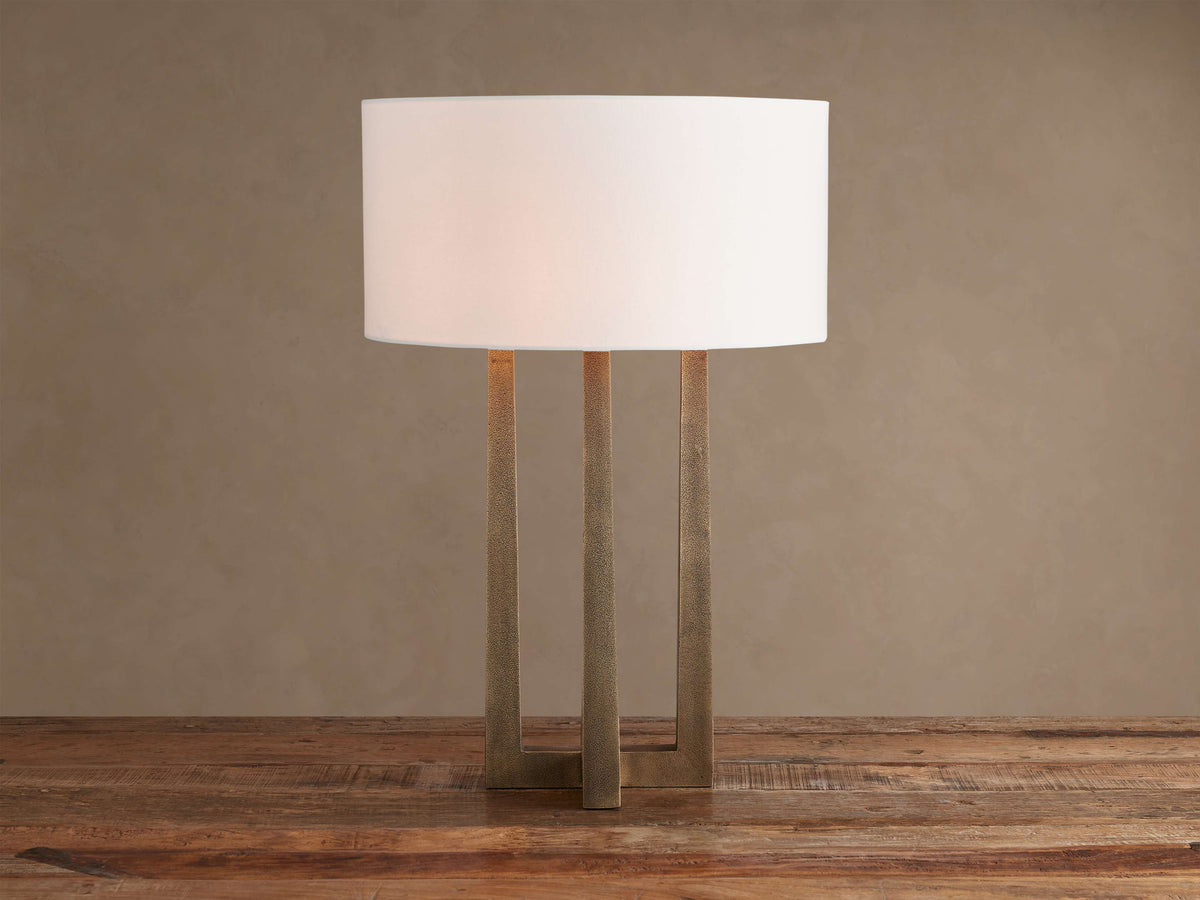 Emmet Table Lamp: lit white cylindrical linen shade, textured antiqued brass three-legged base, on a rustic wood table.